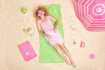 Positive curly haired woman in summer clothes rests on green towel wears headphones on ears enjoys listening music poses at sandy beach surrounded by different items. Summer time and recreation