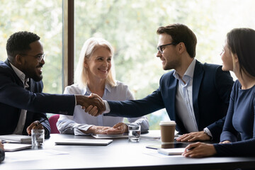 Happy diverse business partners shaking hands over table on partnership meeting, closing deal. Team leader expressing recognition to employee on good work result, achieve, hiring, promotion