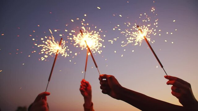 Closeup of hands holding sparklers, happy friends are waving bengal fireworks against background of evening sky. Concept of party