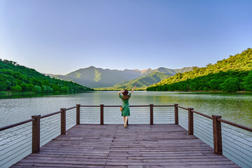 Traveler girl standing alone on edge of pier and staring at lake. Beautiful freedom moment and serene quiet peaceful atmosphere in nature. Back view