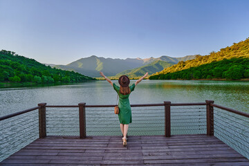 Fototapeta na wymiar Traveler girl with open raised arms standing alone on edge of pier and staring at lake and mountains. Enjoying of beautiful happy free moment life and serene quiet peaceful atmosphere. Back view