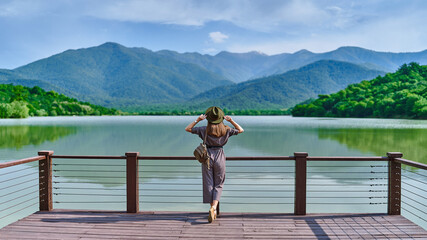Traveler girl standing alone on edge of pier and staring at lake and mountains. Enjoying happy freedom moment life and serene quiet peaceful atmosphere in nature. Back view