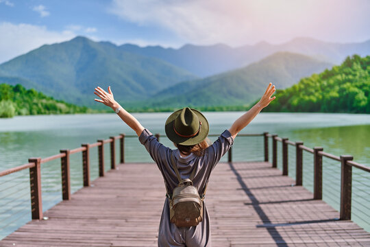 Traveler girl backpacker with open raised arms standing alone on pier and staring at lake and mountains. Enjoying free moment life and serene quiet peaceful atmosphere in nature. Back view