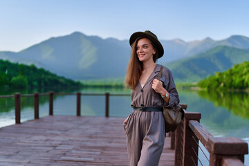 Fototapeta na wymiar Portrait of happy cute smiling attractive wanderer girl wearing hat and backpack standing alone on pier with lake and mountains view. Enjoying serene quiet peaceful atmosphere in nature