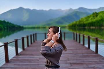 Girl traveler listening to calm music on wireless headphones standing alone on pier with lake and...