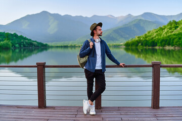 Fototapeta premium Hipster traveler backpacker standing alone on pier with lake and mountains view. Enjoying beautiful freedom moment life and serene quiet peaceful atmosphere in nature