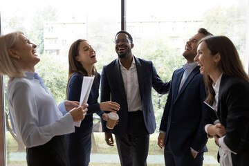 Happy relaxed diverse office colleagues having fun in hallway at window, talking and laughing out loud. Business team discussing funny news, successful project, enjoying work break together