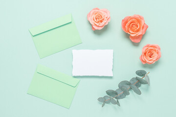 Blank white card mockup with envelopes, roses and eucalyptus branch on mint green background. Wedding stationery template. Top view, flat lay.