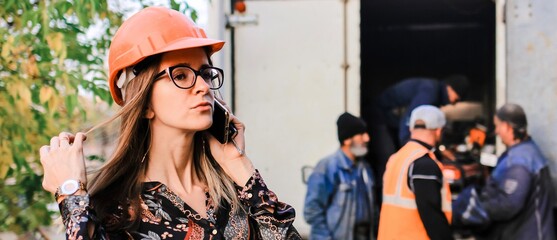 An engineer girl in a safety helmet talking on the phone against the background of construction and workers. Banner.	