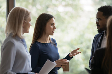 Serious young employee talking to diverse coworkers during office coffee break. Corporate business team standing, chatting in hallway at informal meeting, discussing work, career. Teamwork concept