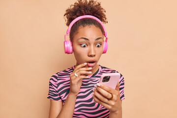 Surprised curly haired ethnic dark skinned woman stares at smartphone display updates playlist wears stereo wireless headphones striped t shirt isolated over beige background reacts on shocking news