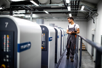 Printing house interior with modern offset print machine and operator in working uniform checking...
