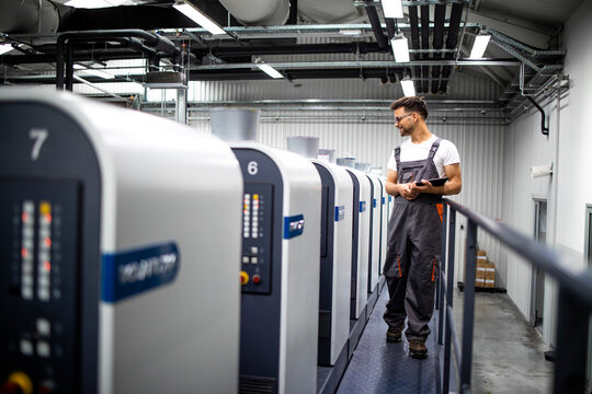 Printing house interior with modern offset print machine and operator in working uniform checking quality and controlling process of print.