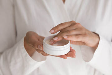 Treat yourself. Close up of millennial female wearing white bathrobe holding cosmetic cream jar. Young lady try moisturizing revitalizing creme lotion on finger before applying on hand face body skin