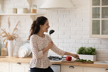 Funny singer. Joyful latin woman vegetarian enjoy easy pleasant cooking of healthy food sing song in microphone of soup ladle. Positive young lady having fun cut vegetables listen to music at kitchen