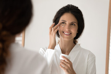 Fresh look every day. Smiling young hispanic woman apply nourishing cream to delicate face skin...