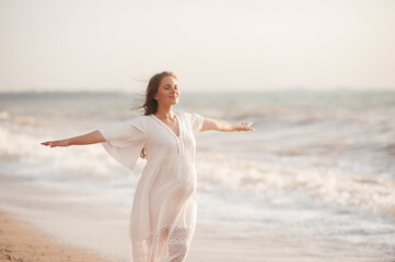 Happy pregnant woman wear white dress feeling free posing over sea background at beach. Motherhood. Maternity. Healthy lifestyle. Carefree adult.
