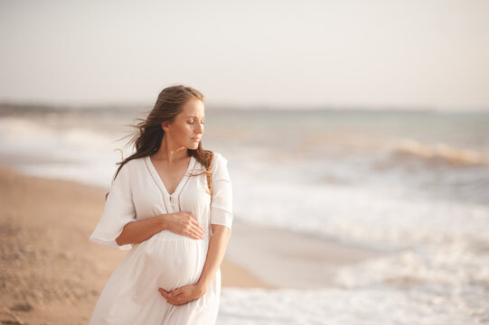 Smiling beautiful pregnant woman wear stylish white dress hold belly walk at beach over waves of sea outdoors. Looking away. Motherhood. Maternity. Healthy lifestyle.