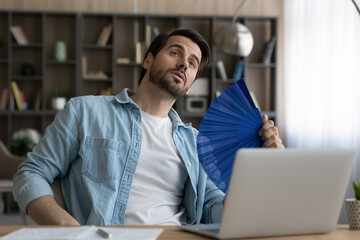 Distracted from computer work anxious unhappy overheated young male worker employee freelancer using paper fan, feeling hot or exhausted in modern home office without air conditioning system.