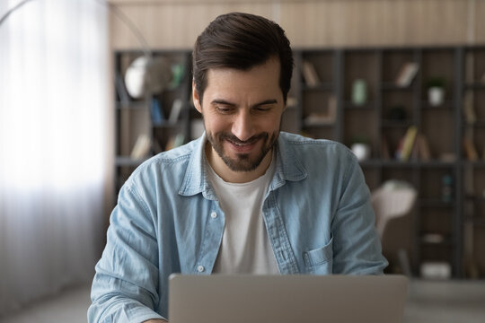 Happy young caucasian man working on computer at modern home office. Smiling millennial guy reading email with pleasant news, enjoying web surfing or shopping in internet store, sitting at table.