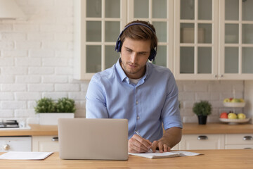Focused millennial man student in wireless headphones learn online use laptop at home kitchen write...