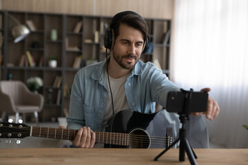 Happy young man in headphones holding guitar, setting up web camera on cellphone, starting...