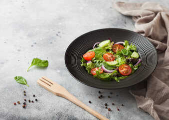 Healthy fresh vegetables salad with lettuce and tomatoes, red onion and spinach in black bowl on dark background with spatula fork and kitchen towel.