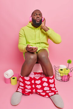 Relaxed dark skinned man with thick beard enjoys listening music via wireless stereo headphones hlds mobile phone sits on toilet bowl in restroom forgets about troubles isolated on pink wall