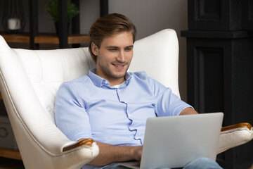 Young bearded man software developer freelancer sit in large soft armchair at home work on laptop...