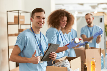 Portrait of happy male volunteer smiling at camera, showing thumbs up and holding tablet pc while sorting, packing food stuff in cardboard box, working on donation project with team. Focus on man