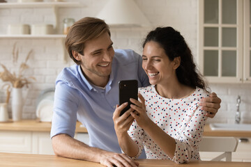 Laughing millennial spouses embrace at home kitchen enjoy watching cute funny video clips photos...