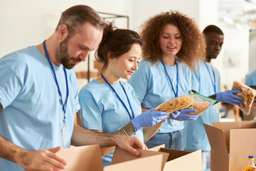 Young woman holding a pack of pasta. Volunteers in protective gloves sorting, packing food in...