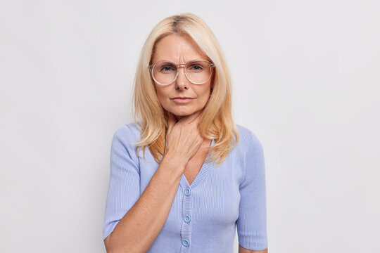 Blonde middle aged woman feels unwell keeps hand on throat difficult to swallow after drinking very cold water wears transparent glasses blue jumper isolated over white background. Health problems
