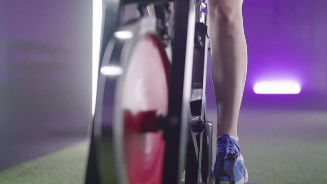Person legs cycling on fitness gym bicycle zoom-out with neon lights 4K