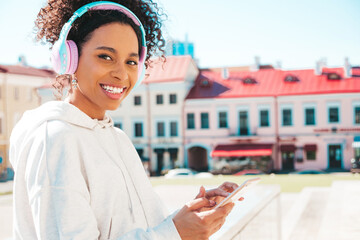 Beautiful black woman with afro curls hairstyle.Smiling model in white hoodie.Sexy carefree female enjoying listening music in wireless headphones.Posing on street background at sunset.Holds phone