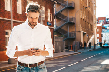 Handsome smiling stylish hipster lambersexual model.Modern man dressed in white shirt. Fashion male posing on the street background. Outdoors at sunset. Using smartphone apps
