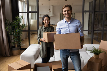Family portrait of happy smiling spouses satisfied real estate agency clients carrying boxes to modern leased purchased apartment. Young couple looking at camera holding packages settling in new home