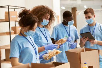 Young team of volunteers in protective masks and gloves sorting, packing food in cardboard boxes,...