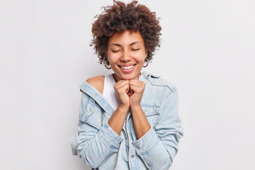 Pretty curly haired ethnic woman closes eyes smiles broadly keeps hands under chin has gentle expression wears denim jacket isolated over white background feels heartwarming stands romantic.