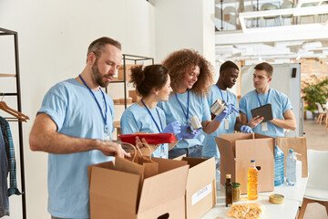 Team of diverse volunteers in protective gloves sorting, packing foodstuff in cardboard boxes,...