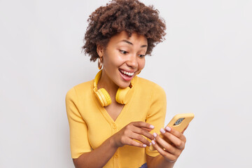 Smiling adorable teenage girl with curly hair chats online via smartphone uses application addicted to modern technologies wears stereo headphones around neck casual yellow jumper white background