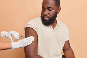 Young bearded ethnic man with dark skin gets injection in arm to stop coronavirus poses against...