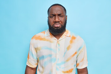 Discontent dark skinned man with thick beard has displeased sulking expression washed out t shirt discontent by something isolated over blue background. Negative huan emotions and face expressions