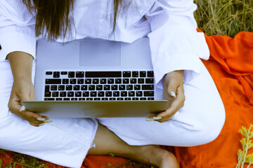 An unrecognizable girl in a white suit is typing on a laptop keyboard sitting on an orange plaid in nature. Yellow flowers lie on the computer. Online business, communication, business, work concept.