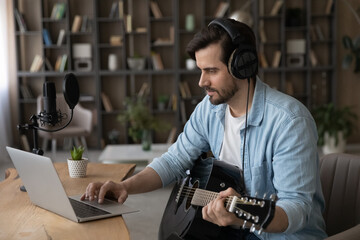Motivated smiling handsome young man web surfing educational music video, practicing playing on guitar at home, improving skills alone indoors. Happy musician recording own song using laptop.