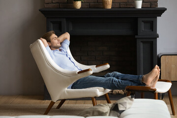 Carefree young male homeowner relaxing barefoot in capacious armchair at luxury apartment by modern fireplace. Serene single millennial male rest with hands behind head at rich comfortable living room - obrazy, fototapety, plakaty