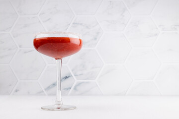 Fresh strawberry margarita cocktail in high glass goblet with sugar rim in soft light white kithchen interior witj marblr tile.