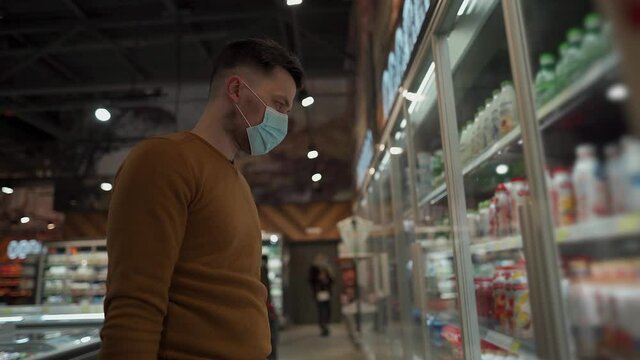 Young Caucasian Man In Face Mask Choosing Dairy Products At Supermarket During Coronavirus Pandemic. Male Shopping At Grocery Store Wearing Facemask, Shopping Basket In Hand. Topic New Norm Covid 19.