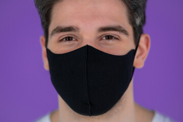 Portrait of handsome man in facial medical mask looking to camera. Guy in studio on bright purple background. Covid-19, coronavirus, new reality concept.