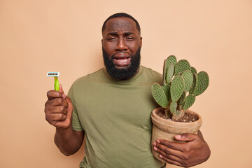 Unhappy crying dark skinned ethnic man holds razor and potted cactus has thick stubble as prickly...